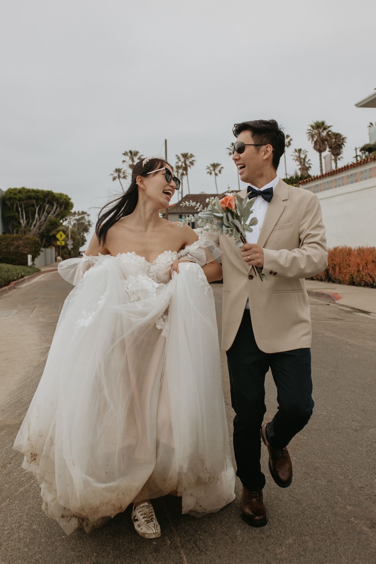 A couple in wedding attire run together on a palm tree-lined street on a cloudy day at Windansea Beach in La Jolla.