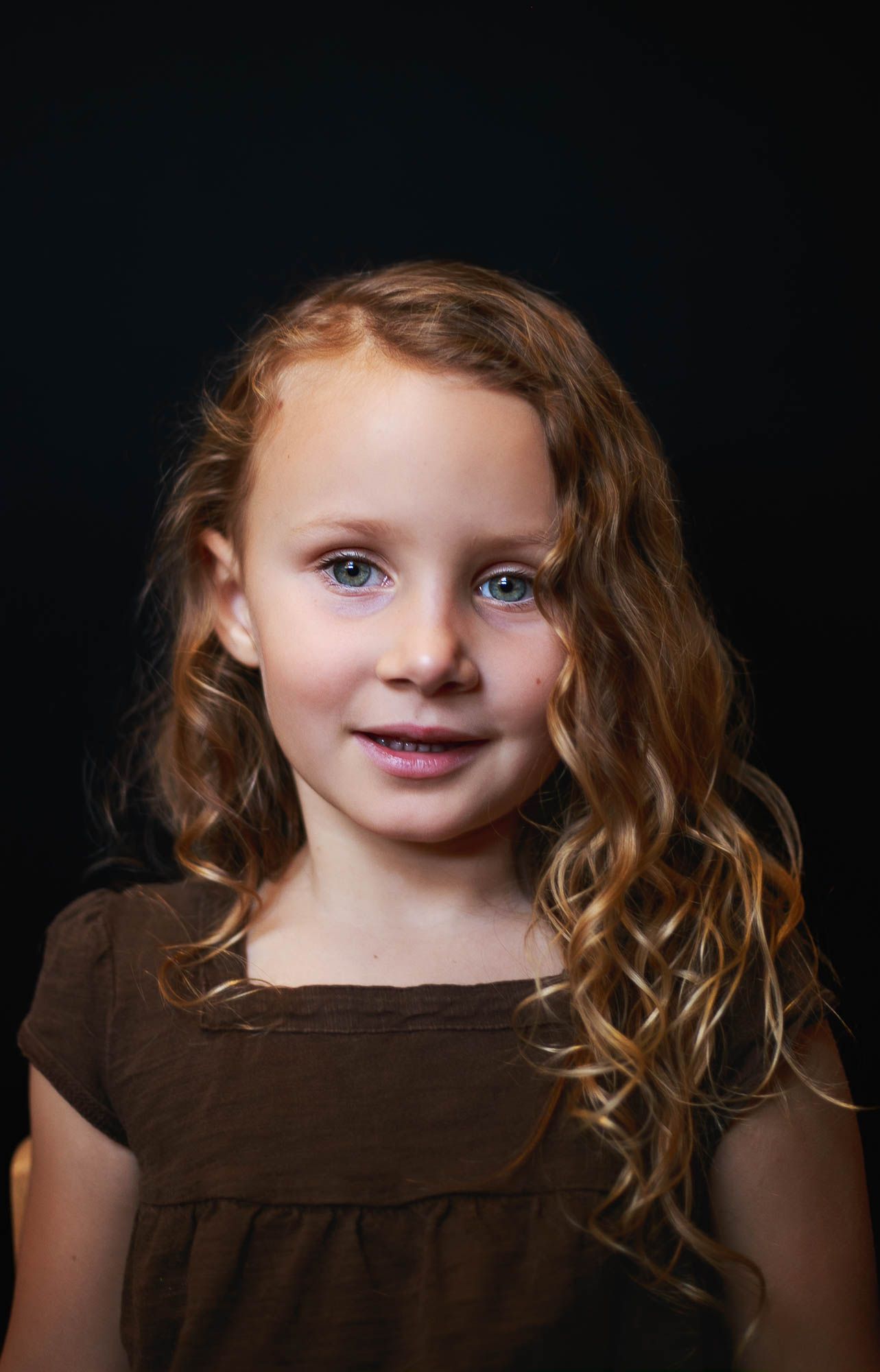 A fine art portrait showing a child with curly hair wearing a brown shirt against a dark background.