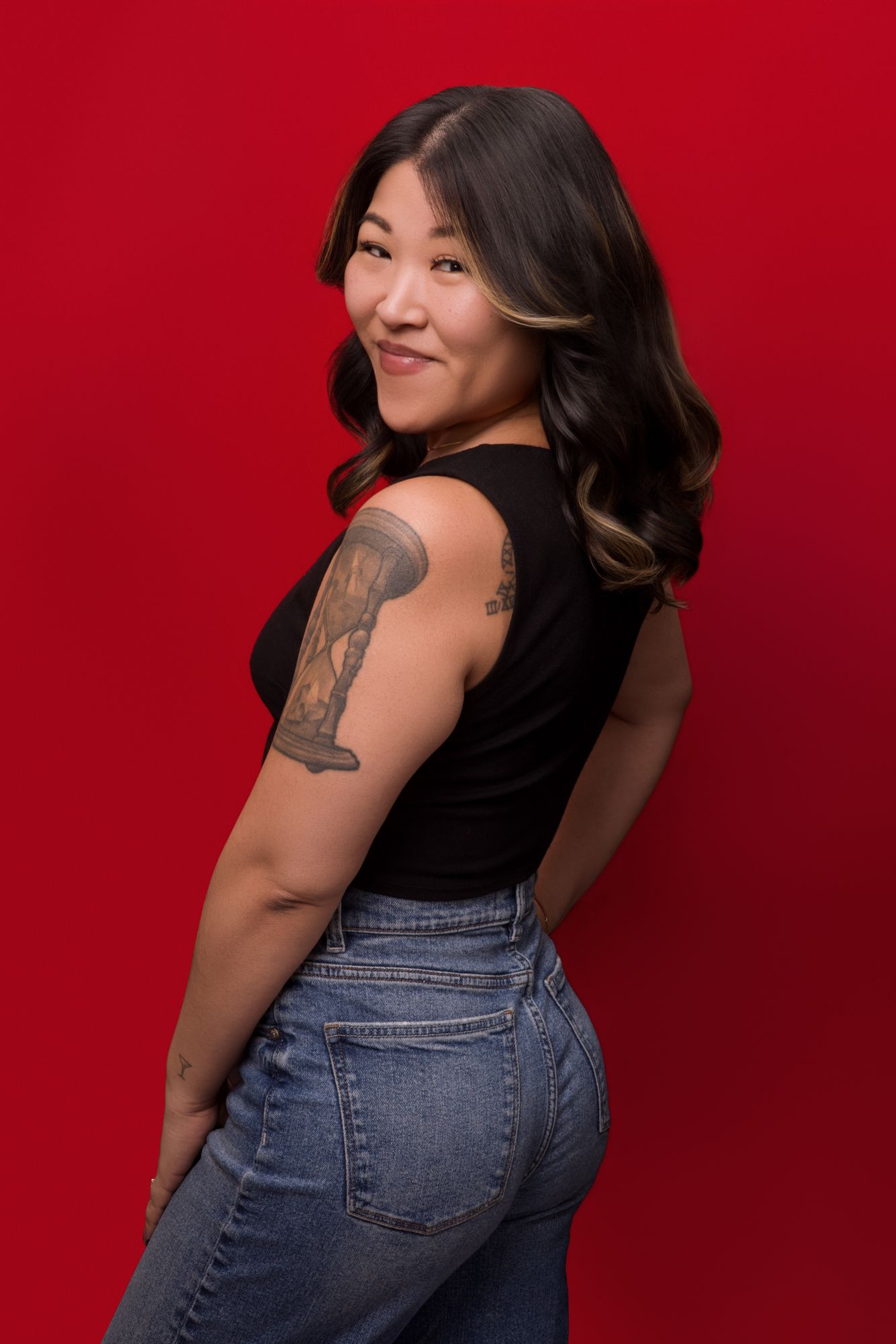 A person in a black tank top and jeans poses against a red background, showing off a detailed arm tattoo.