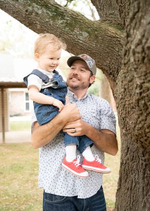 A person in a patterned shirt holds a small child near a large tree trunk in an outdoor setting.