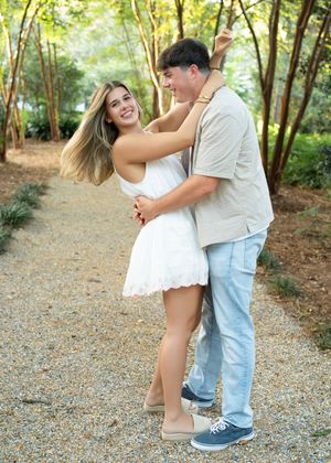 A couple dances playfully together on a gravel path surrounded by lush green trees and forest scenery.