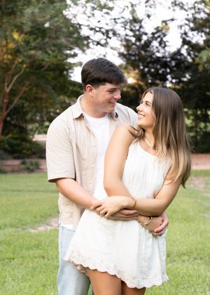 A couple embraces while standing on grass in a garden setting, sharing a tender moment together.