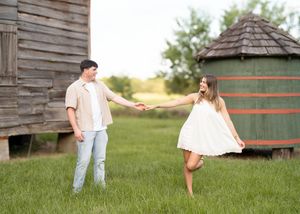 A couple shares a playful moment together on a farm with rustic wooden buildings and a silo in the background.