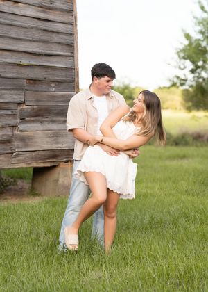 A couple dances playfully together in a grassy field next to a rustic wooden barn wall.
