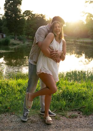 A couple embraces by a serene lake at sunset, standing on a dirt path surrounded by lush green grass.