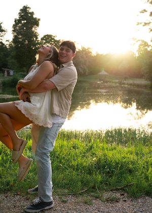 Couple embraces in romantic dance pose by a tranquil lake at sunset with lush greenery in the foreground.