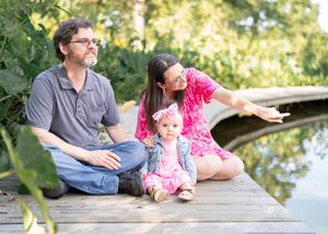 A family sits together on a wooden dock by a pond on a sunny day surrounded by lush greenery.