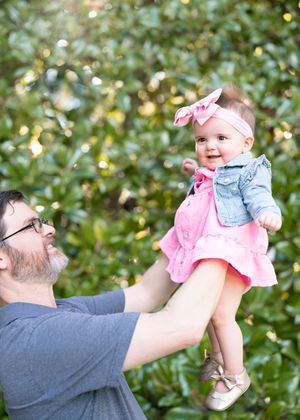 A baby wearing a pink dress and denim jacket is lifted up against a leafy green background.