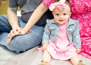 Baby wearing pink dress, denim jacket and bow headband sits with family members on ground outdoors.
