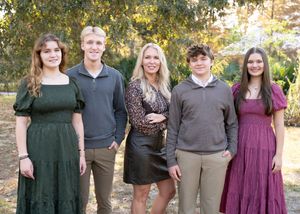 A group of people in coordinated formal attire pose together outdoors against a backdrop of autumn trees.
