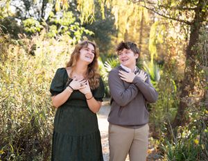 Two people share joyful laughter while standing together in a sunlit autumn forest setting.