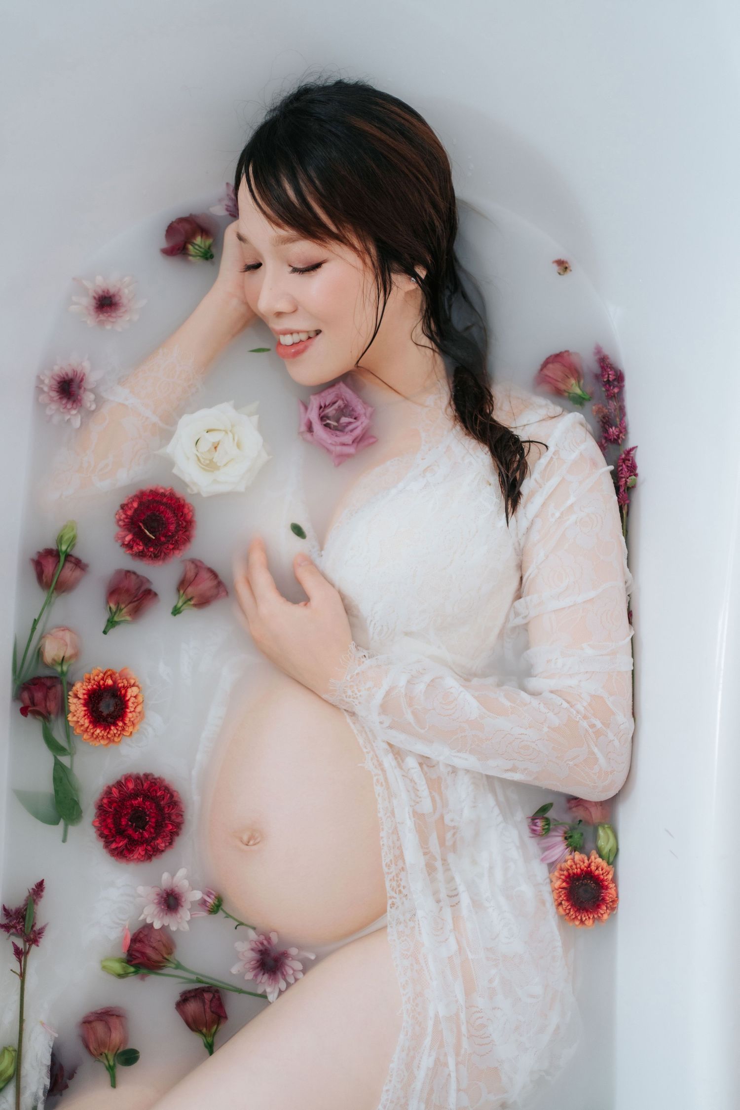 Smiling pregnant woman in white lace robe relaxing in a milk bath surrounded by colorful flowers.一位微笑的孕婦身穿白色蕾絲睡袍，在牛奶浴中放鬆身心，周圍環繞著五彩繽紛的鮮花。