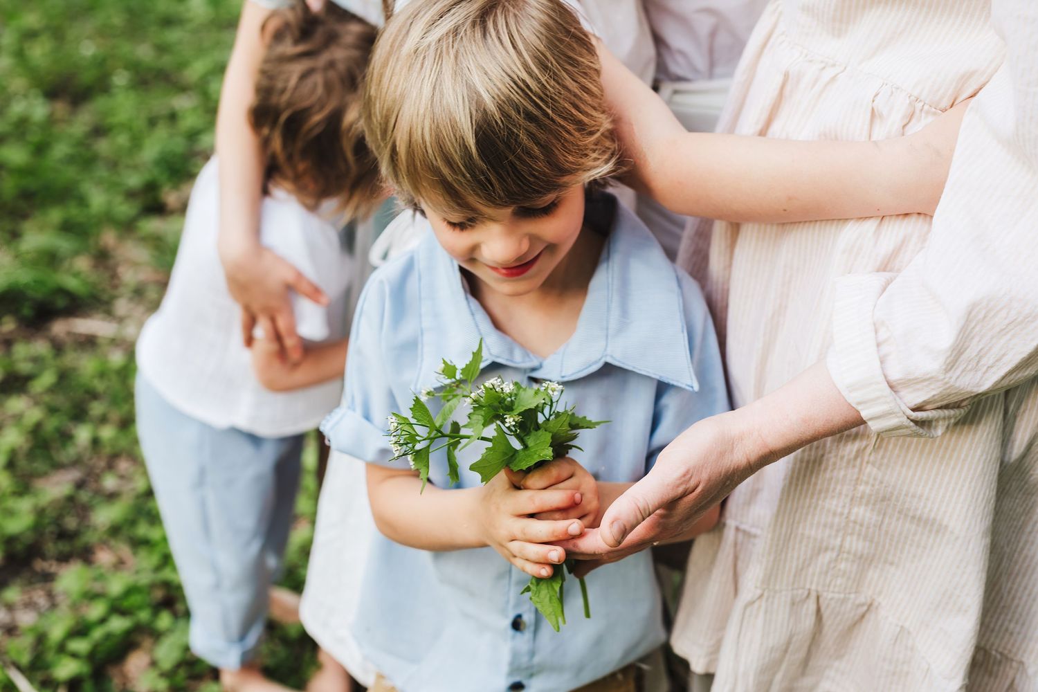 Child in light blue shirt holds fresh herbs while learning about gardening with adult in outdoor setting.