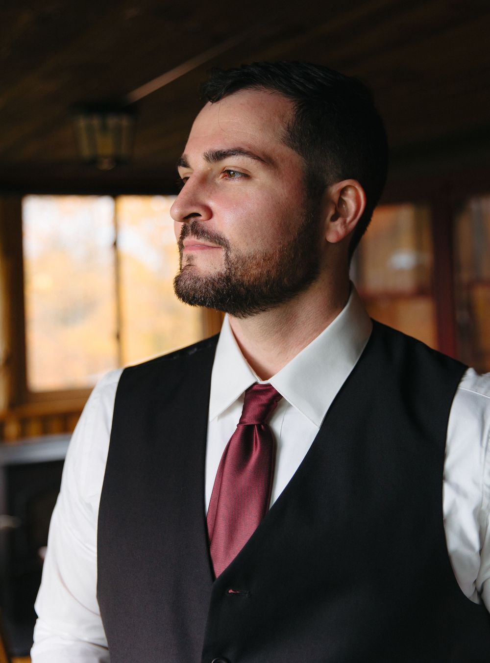 Formal portrait of person in vest and burgundy tie looking to the side in warm lighting.