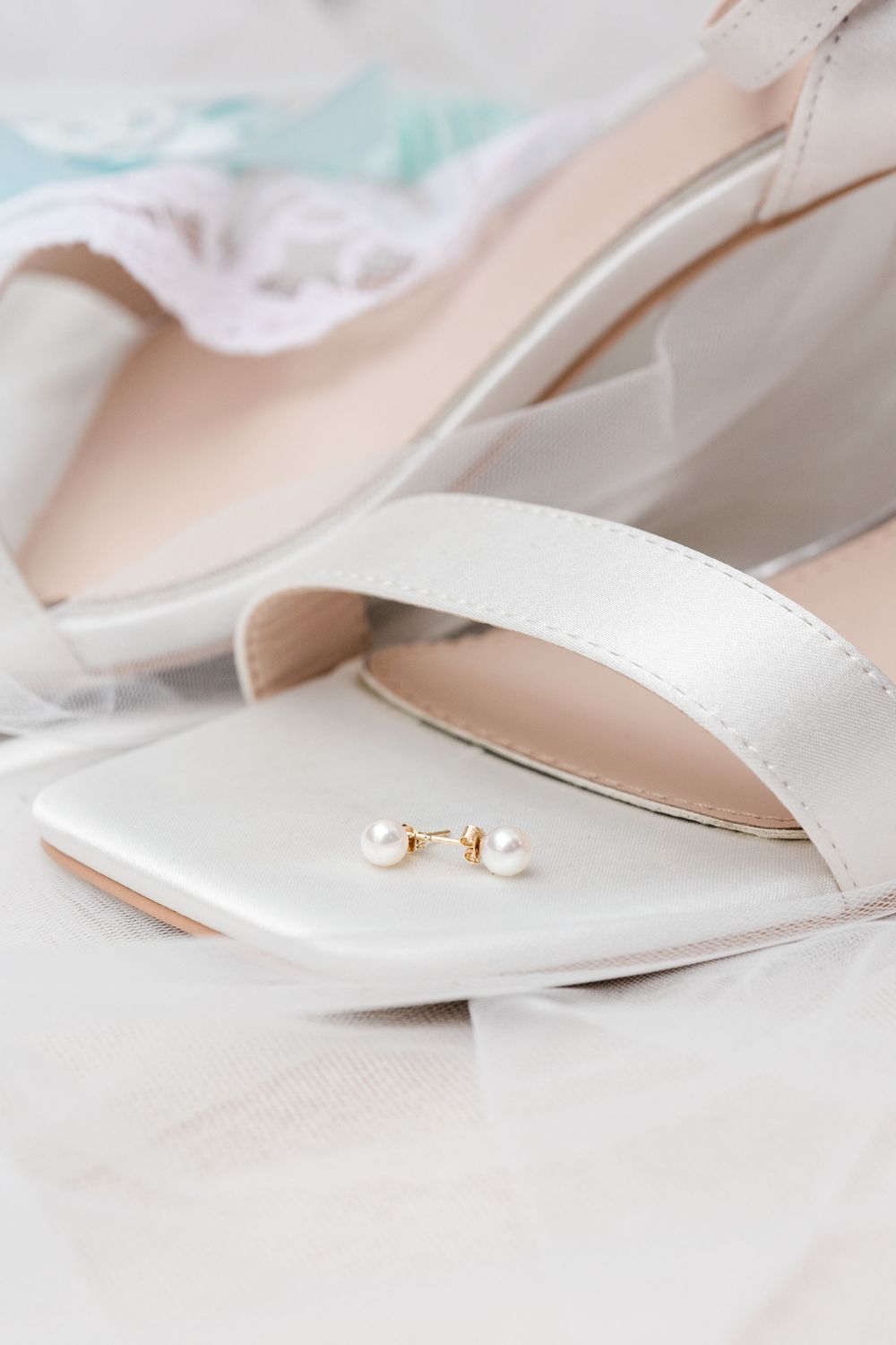Pearl earrings resting on elegant white bridal shoes with soft fabric in the background.