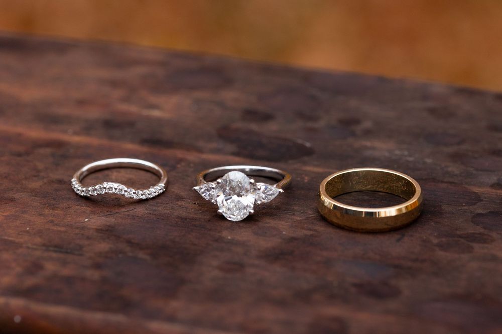Three wedding rings displayed on a dark wooden surface, including a diamond engagement ring and two wedding bands.