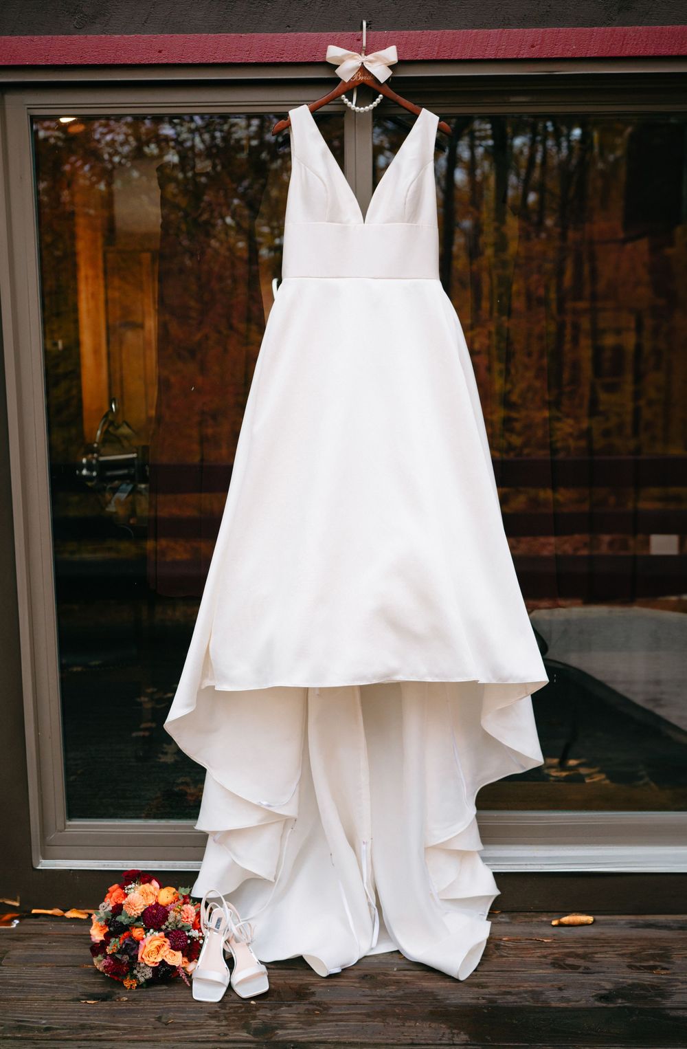 Elegant white wedding dress with v-neck and train hanging against glass doors with autumn trees behind.