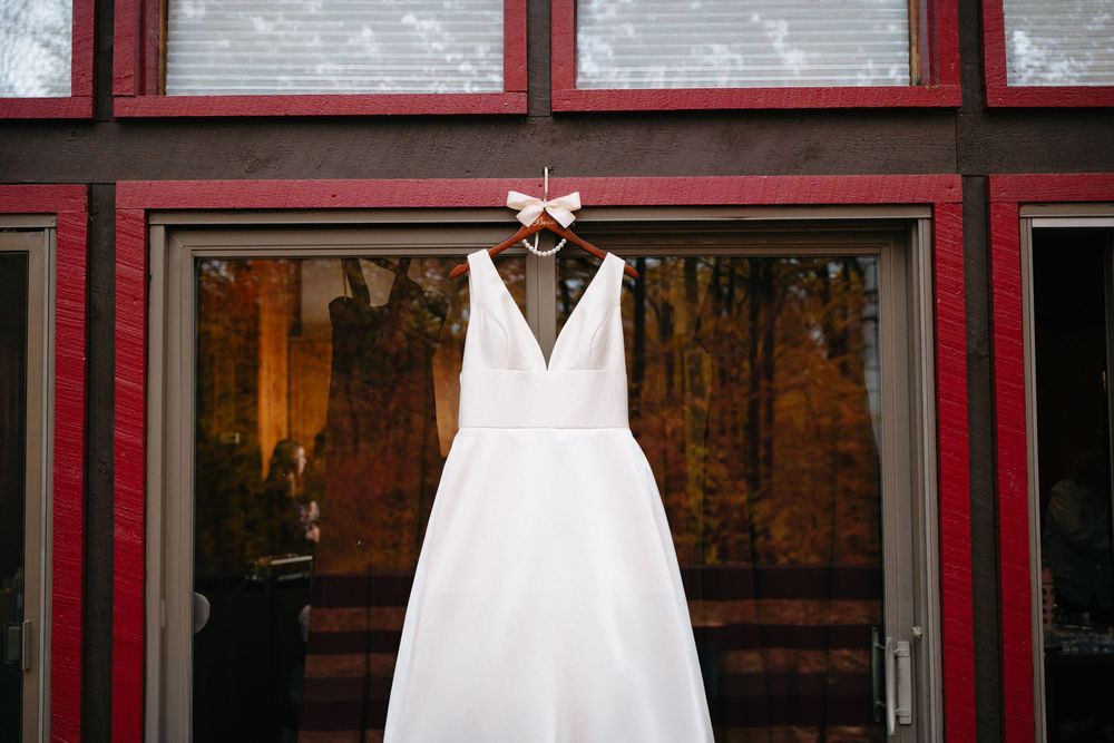 A white wedding dress hangs from a hanger against red-framed windows with a rustic wooden backdrop.