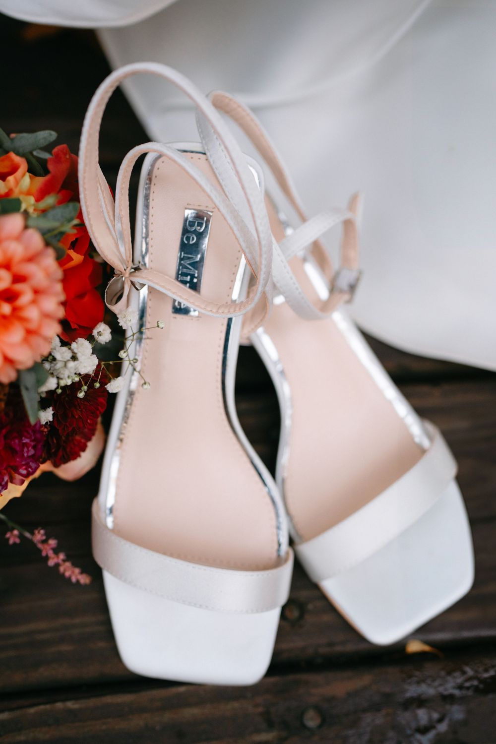 White slingback sandals with block heels sit beside coral flowers on a dark wooden surface.