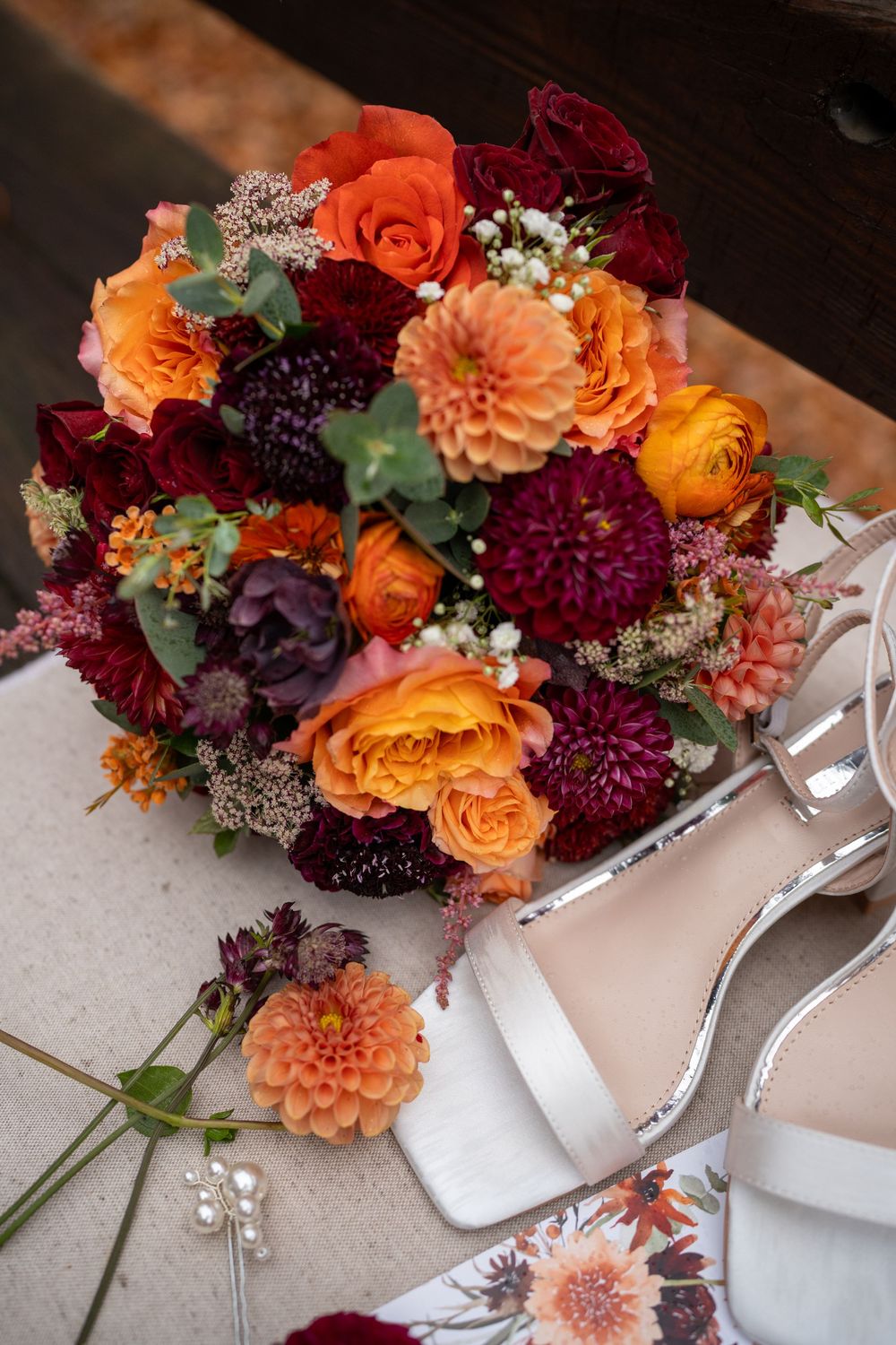 Vibrant fall wedding bouquet with orange roses, burgundy dahlias and white flowers next to bridal shoes.