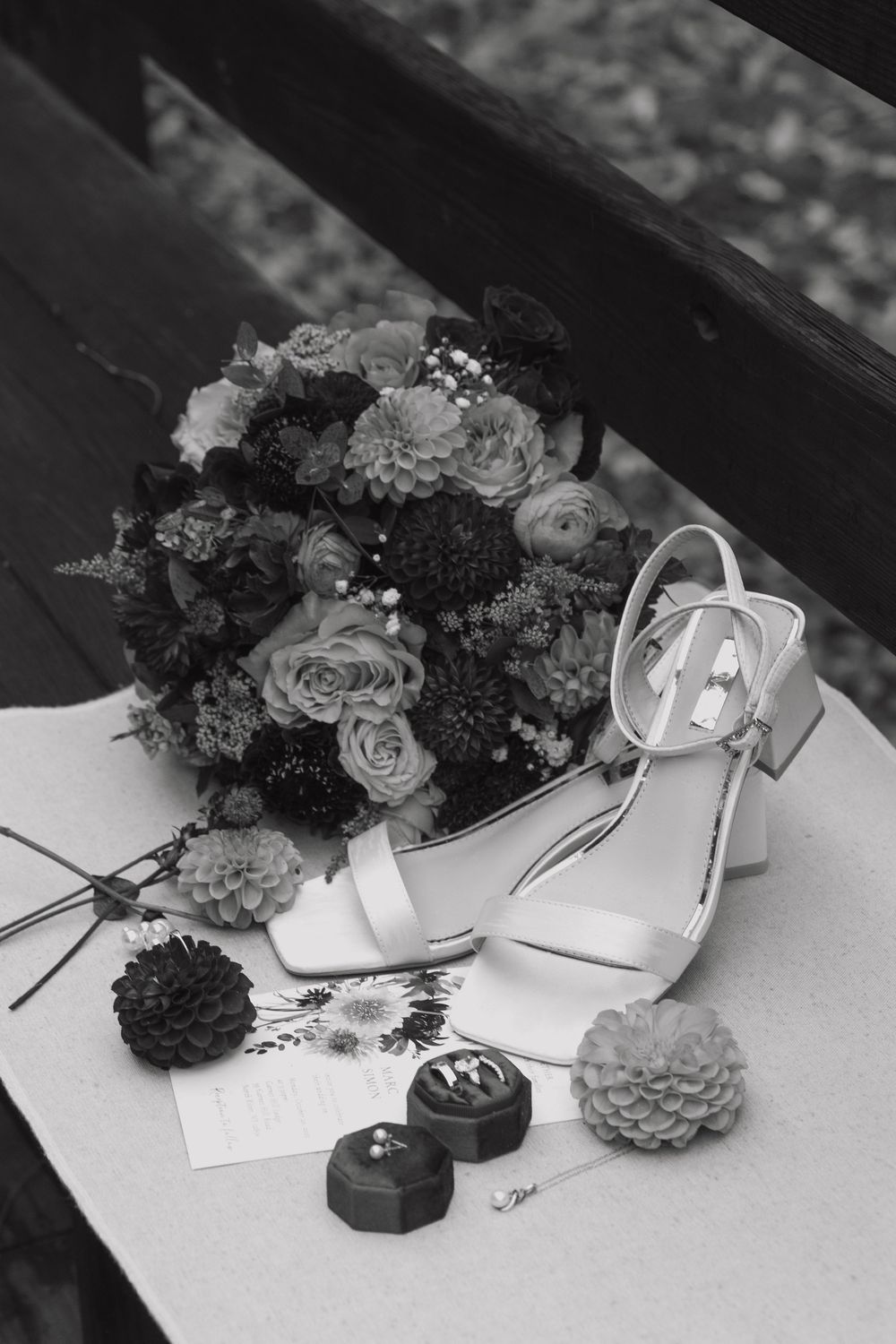 Black and white photo of wedding shoes and floral arrangement with chocolates on white surface.
