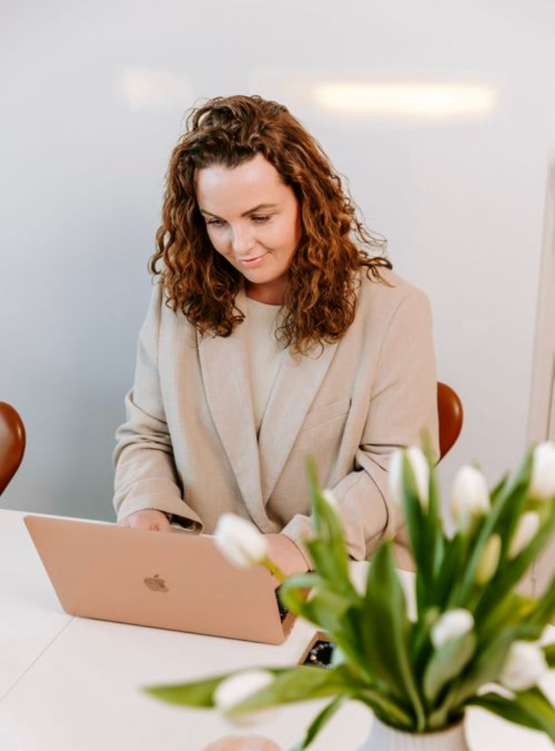 En professionel i en beige blazer arbejder på en MacBook med hvide tulipaner i forgrunden på en moderne arbejdsplads. Branding fotograf Fyn.