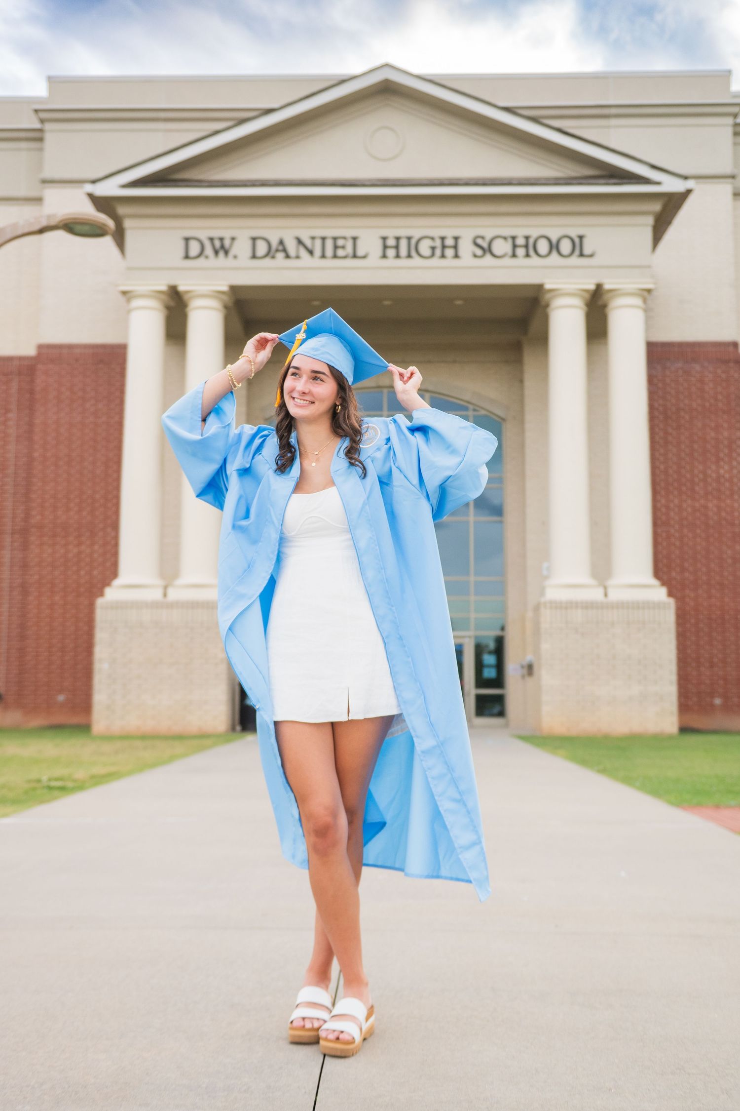 Graduate in light blue cap and gown poses in front of D.W. Daniel High School entrance on graduation day.