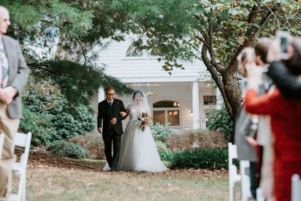 A wedding ceremony takes place in a serene garden setting with lush greenery and a white house in the background.
