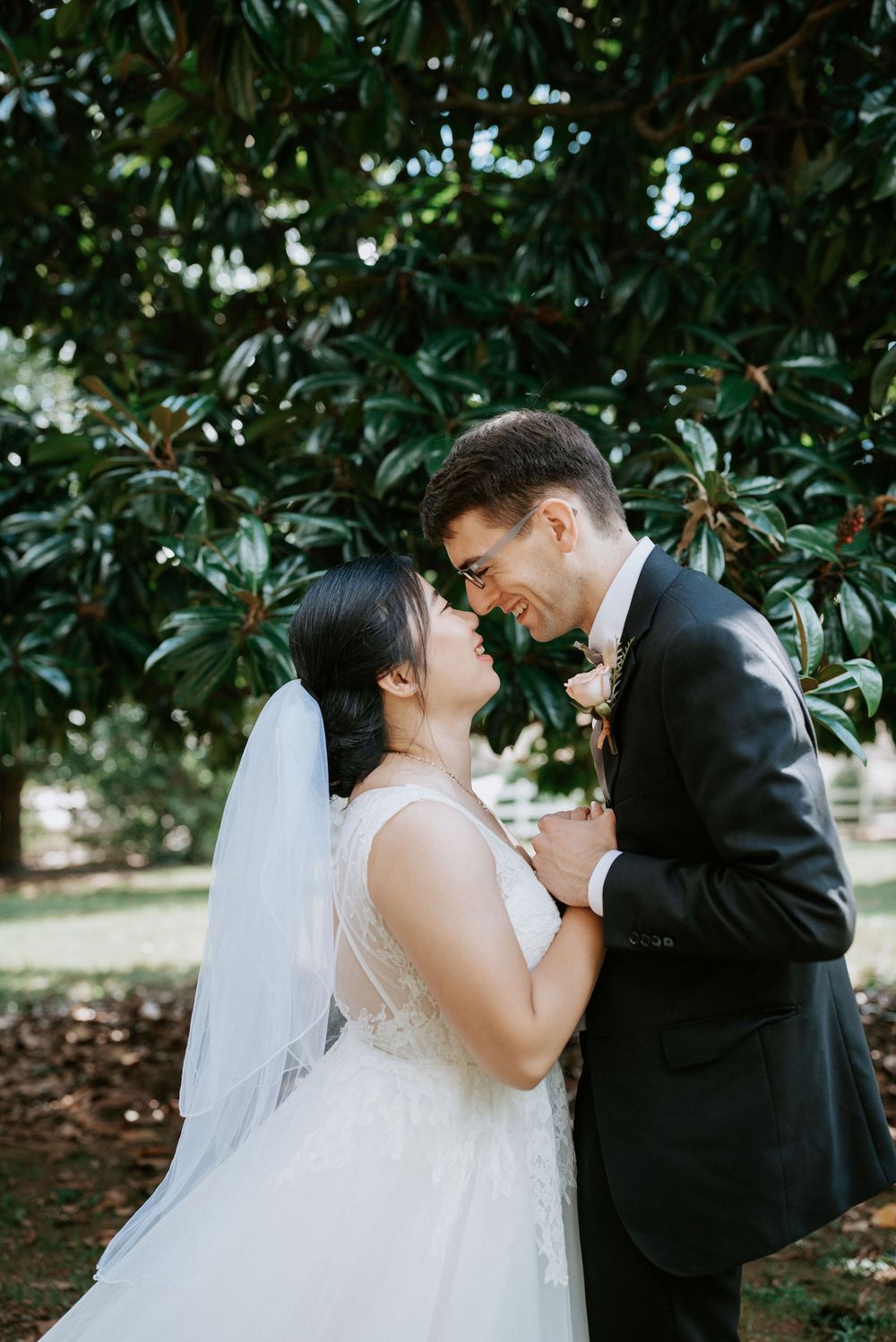A romantic wedding couple shares tender moments under lush green trees in a garden setting.