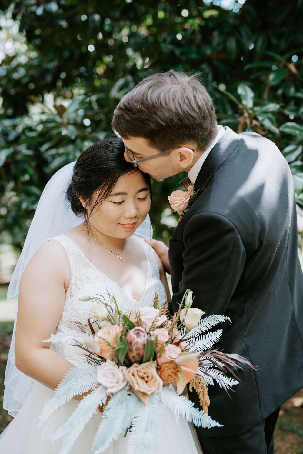 A tender wedding moment outdoors with a beautiful bouquet featuring pink roses and pampas grass against greenery.