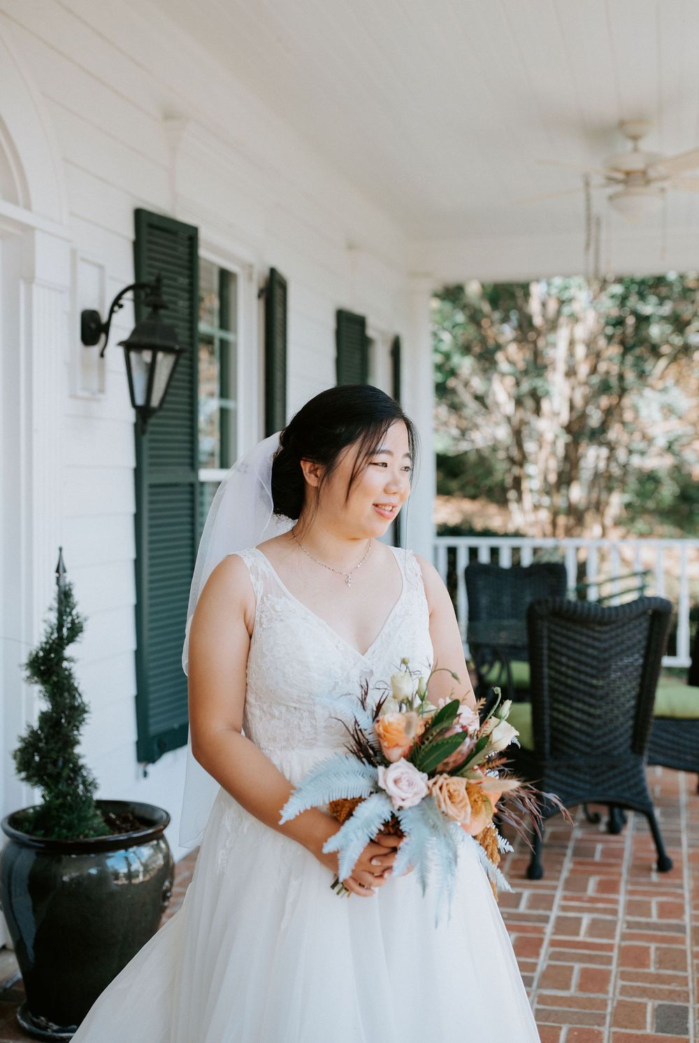 A bride in a white lace gown holds a peach and white floral bouquet while standing on a brick porch with green shutters.