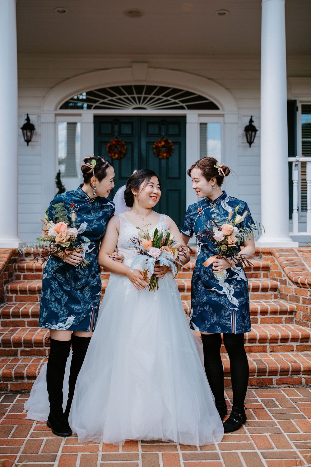 Wedding party wearing navy blue dresses and white gowns pose on brick steps in front of a white columned porch.