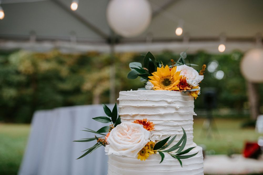 A rustic white wedding cake decorated with sunflowers and roses sits under glowing lanterns at an outdoor reception.