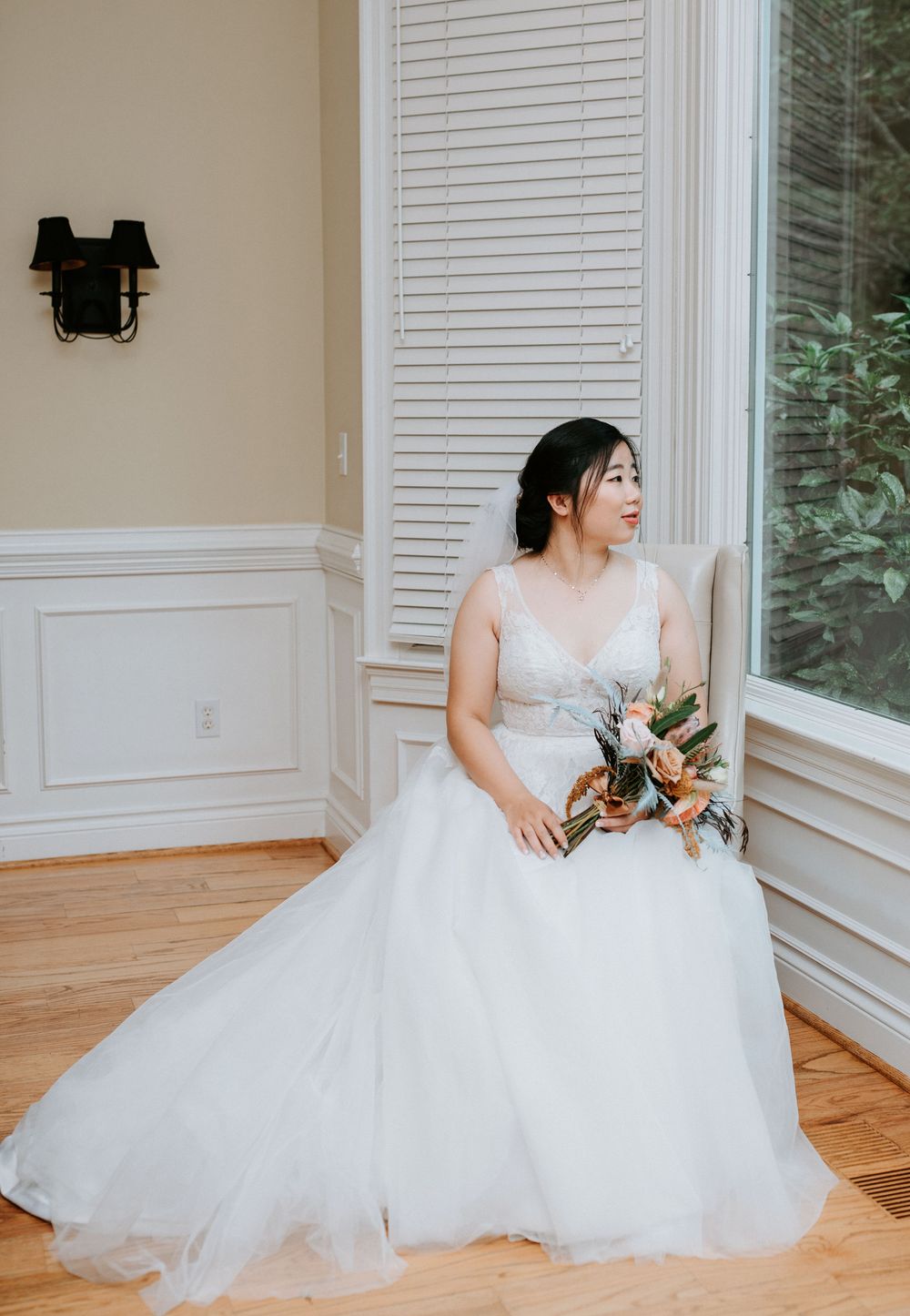 A bride in a flowing white tulle wedding gown sits gracefully on a hardwood floor near large windows with white shutters.