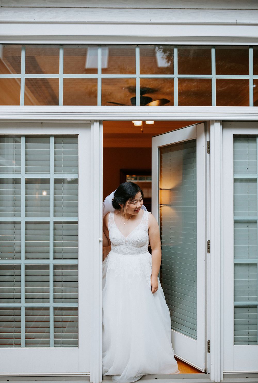 A bride in a white lace wedding dress stands in an elegant doorway with transom windows above.