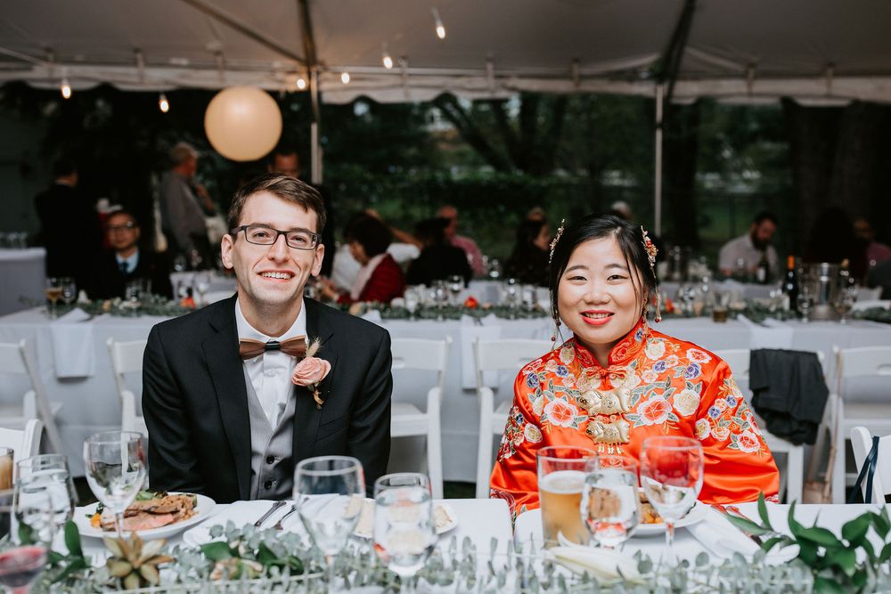 A couple sits at an elegant outdoor wedding reception table under a white tent with paper lanterns.
