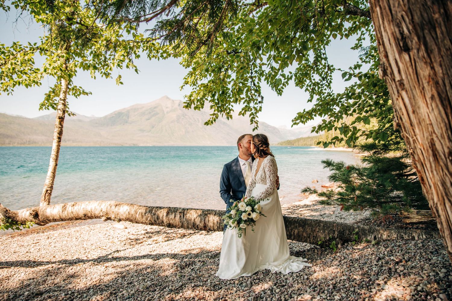 Couple shares a romantic moment under tall trees along a scenic lakeside beach with mountains in the background.