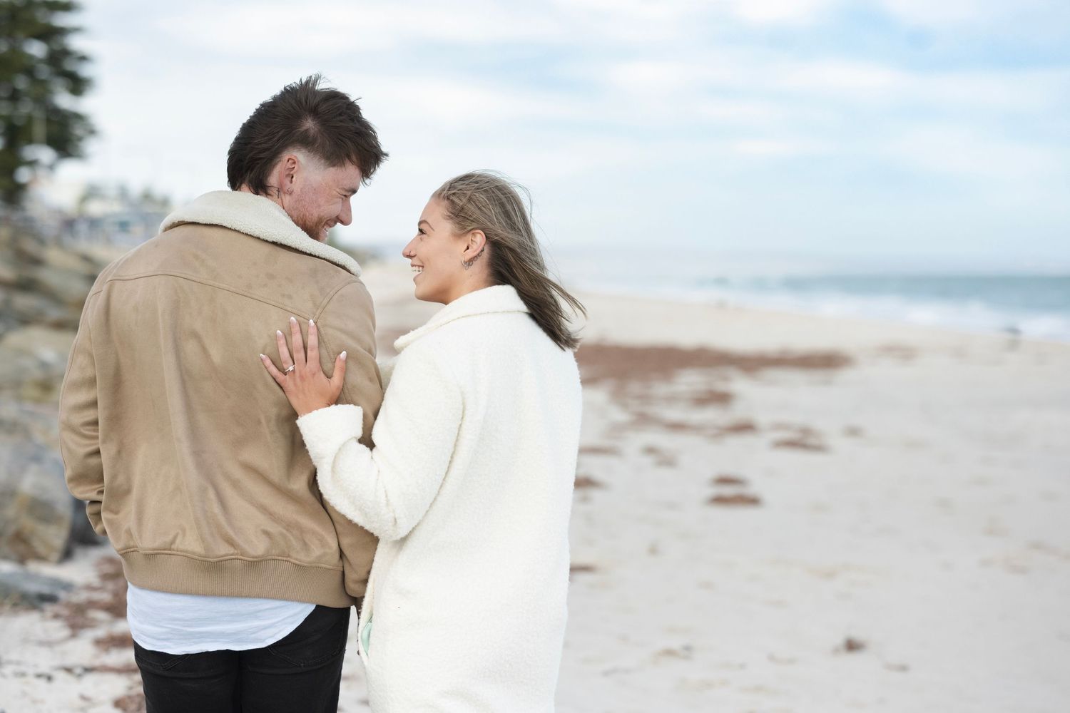 Romantic couple embracing on a sandy beach, sharing a loving gaze while dressed in casual winter outfits.