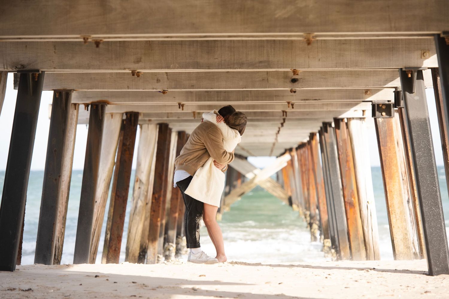 Couple embracing romantically under a wooden pier on a sandy beach with ocean waves in the background.