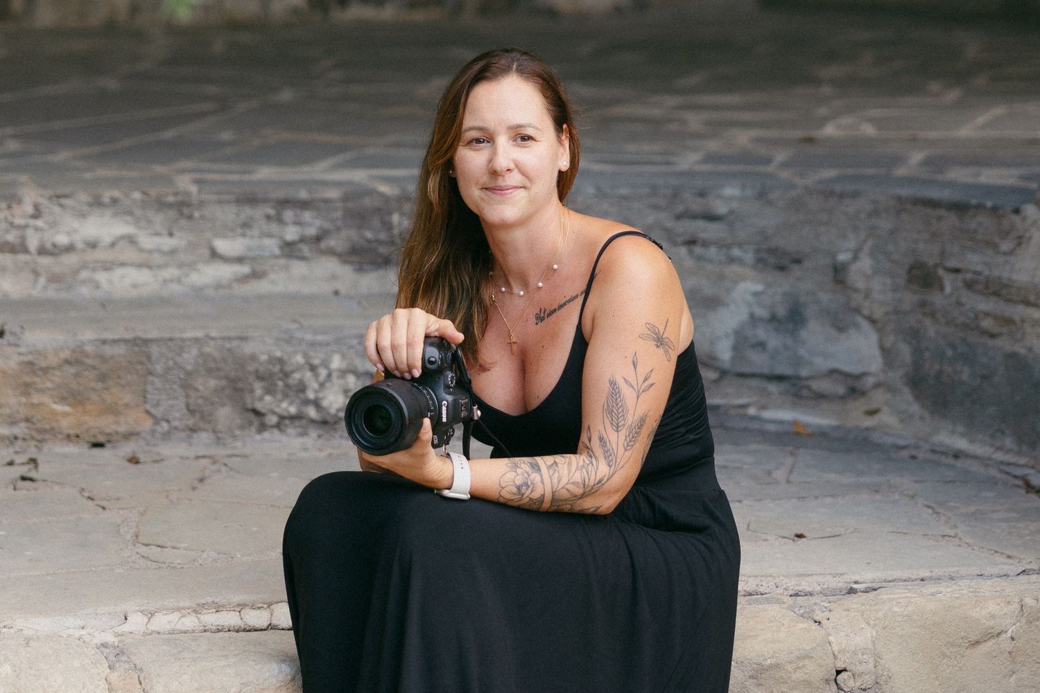 Professional photographer in black dress holds camera while sitting on rocky surface outdoors in Paphos.