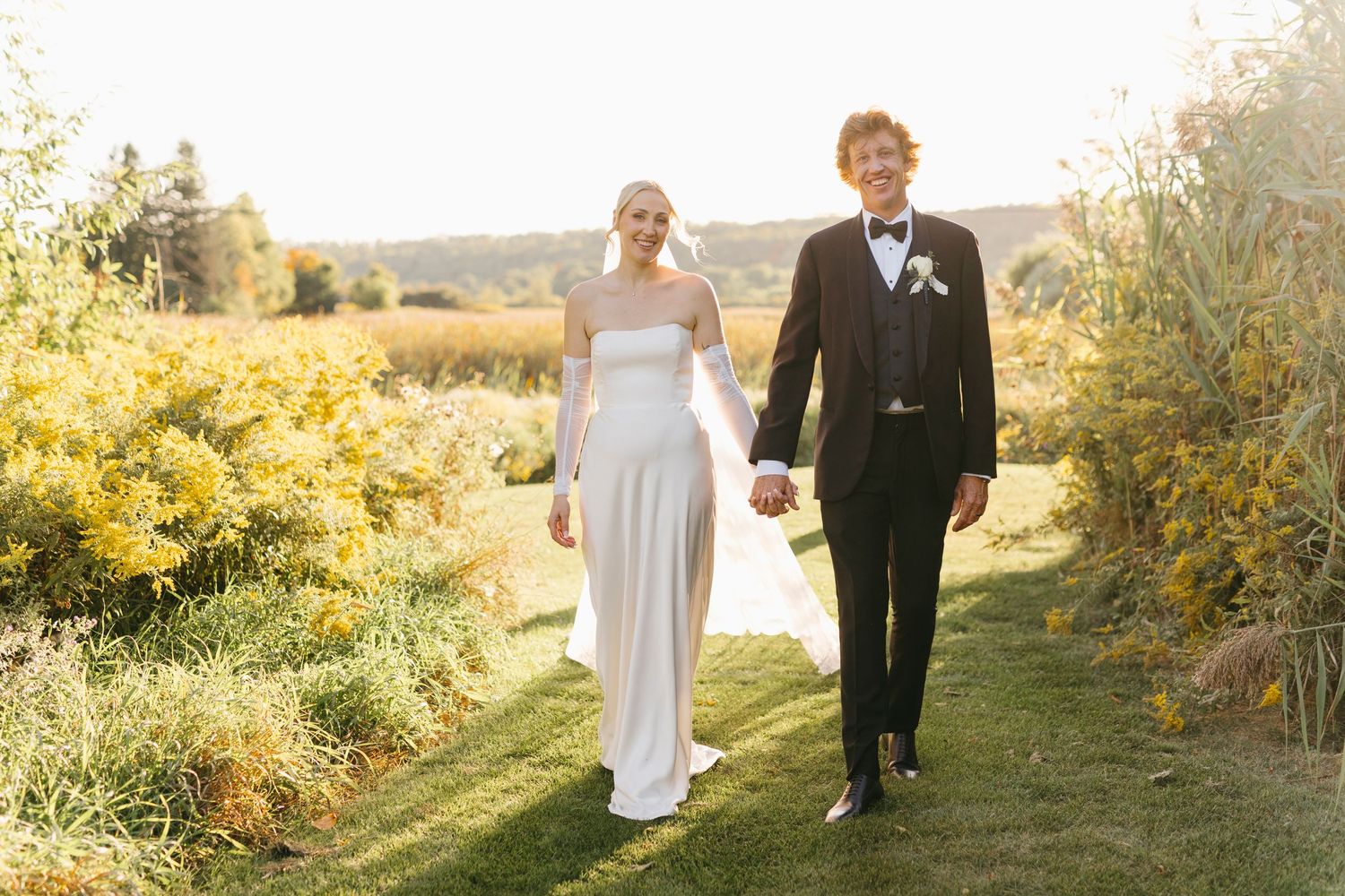 Bride in strapless white gown and groom in black tuxedo walk through autumn countryside landscape.