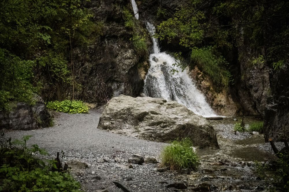 A cascading waterfall flows down rocky cliffs surrounded by lush green forest in a moody natural setting.