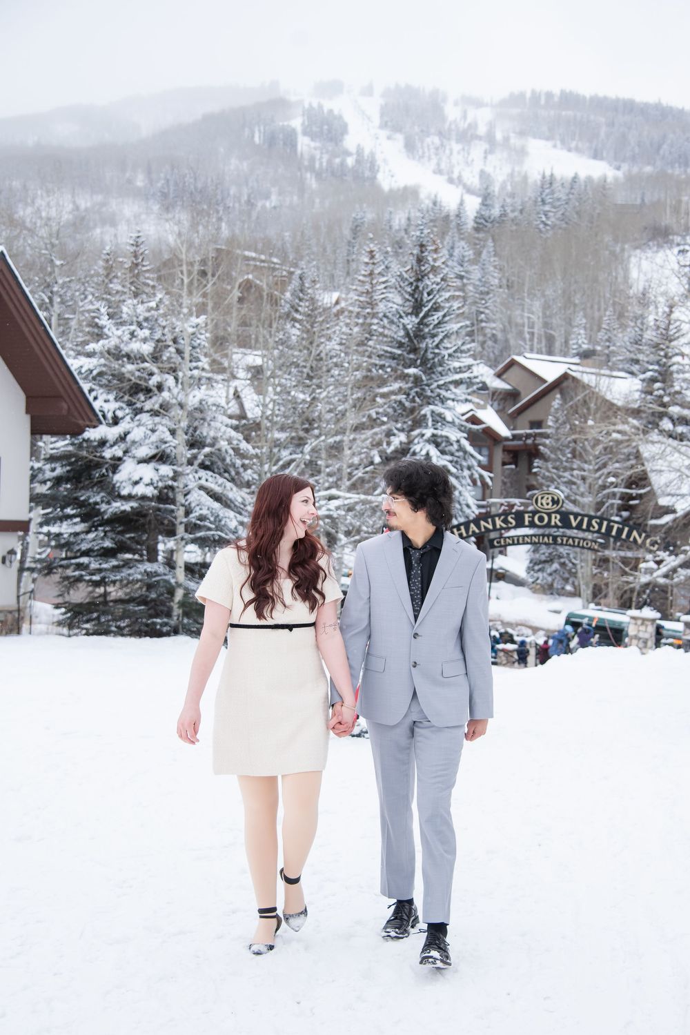 A couple walks hand in hand through a snowy winter landscape with ski slopes and mountain lodges in the background.