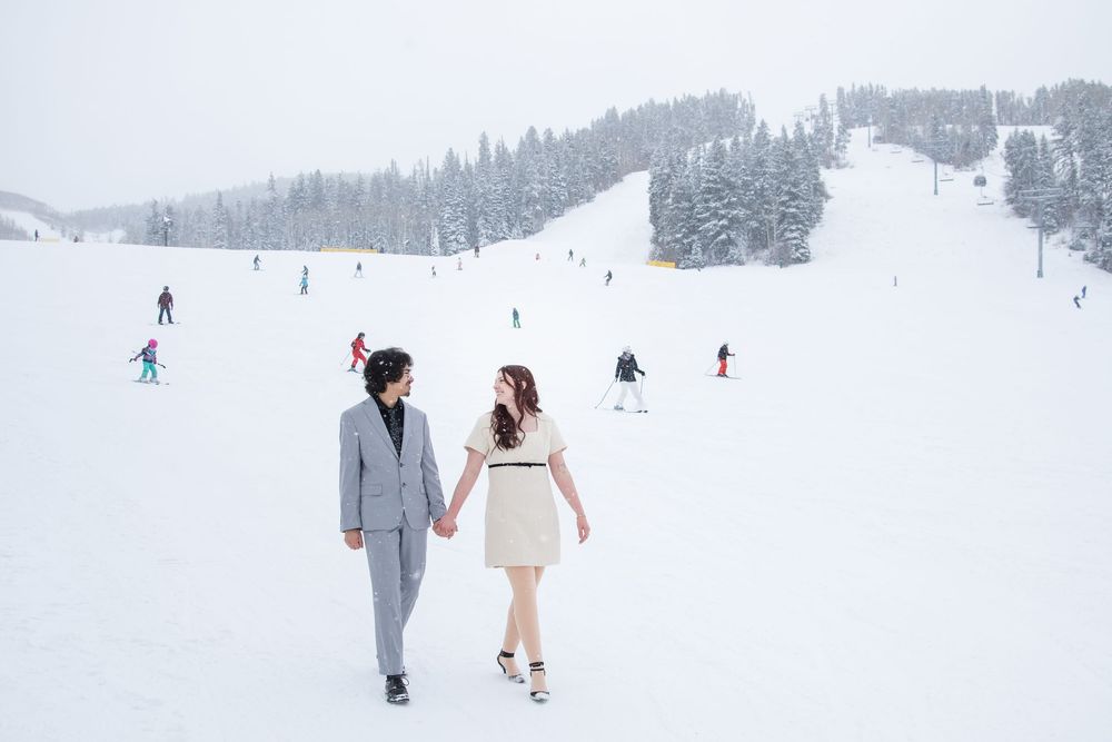 A couple walks together in winter wedding attire through a snowy mountain landscape with skiers in the background.