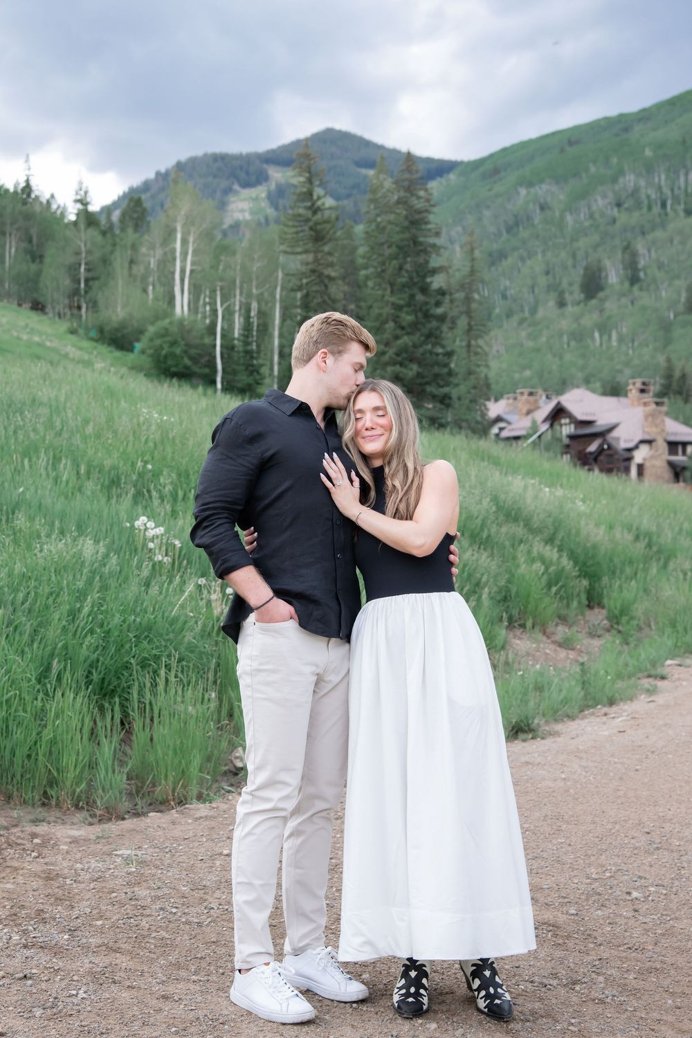 A couple embraces on a mountain trail surrounded by evergreen trees and rolling hills in the background.