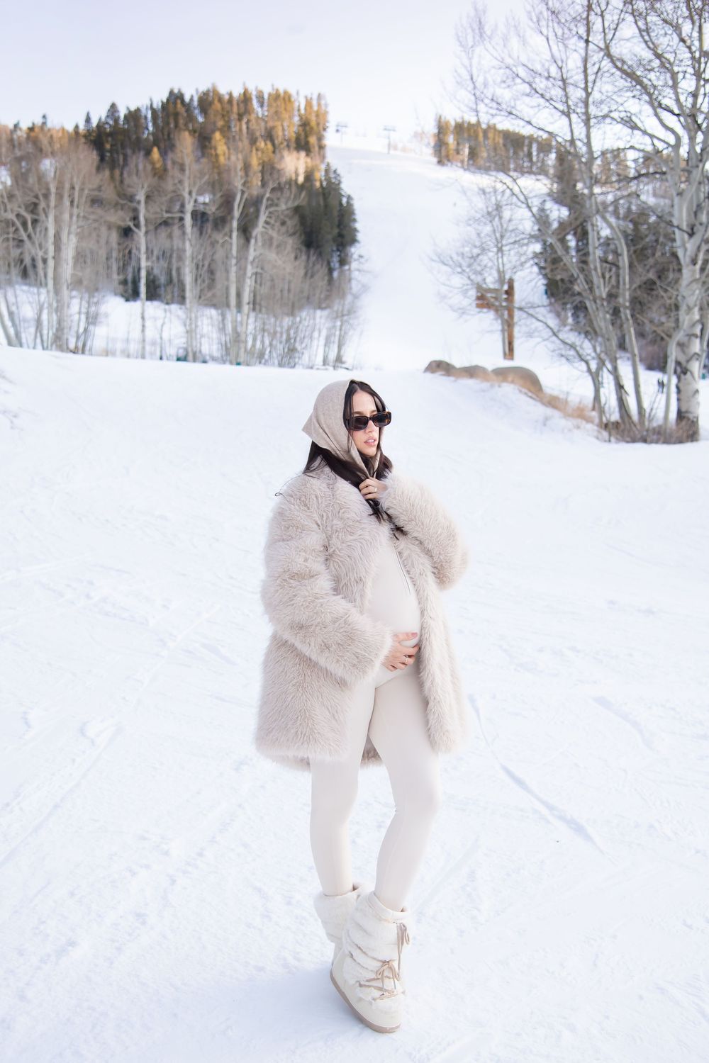 A figure in a plush cream-colored snowsuit poses playfully in a snowy winter landscape with bare trees in the background.
