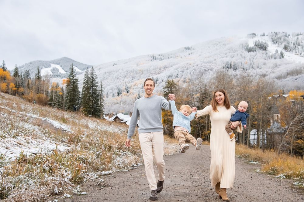 A couple walks hand in hand through a snowy mountain landscape with evergreen trees in the background.