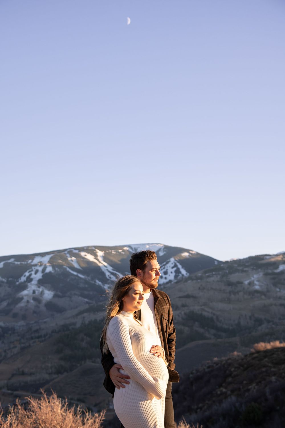 A couple embraces against a scenic mountain backdrop with snow-capped peaks during golden hour.
