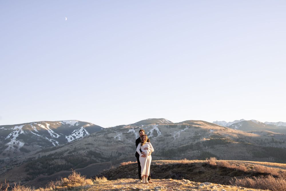 A couple stands on a mountain summit during golden hour with snow-capped peaks in the background.