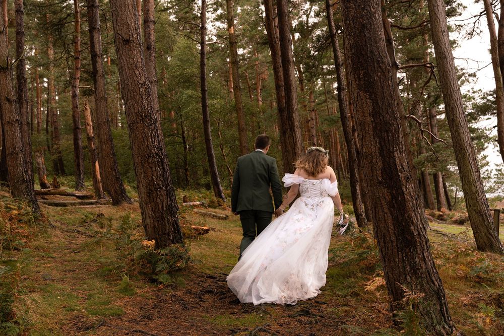 Couple in wedding attire walk hand in hand through a serene pine forest path.